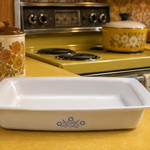 May include: A rectangular white ceramic baking dish with a smooth, glossy finish. The front exterior features a small, centered blue floral pattern consisting of stylized flowers and leaves. The dish has a wide, flat rim and deep, straight sides.