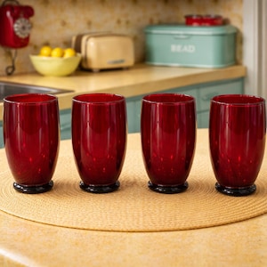 May include: Four ruby red drinking glasses with black bases are arranged on a round, woven placemat. The glasses are cylindrical with a slight taper and are set on a light-colored countertop in a kitchen setting.