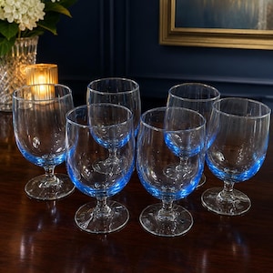May include: A set of six clear glass goblets with a distinct blue tint at the base of each bowl. The glasses feature a rounded bowl shape, a short stem, and a circular foot, providing a consistent design across the entire collection of drinkware.