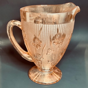 May include: A vintage pink glass pitcher with a handle and a spout. The pitcher has a ribbed texture and is decorated with floral designs. The pitcher sits on a pedestal base. A classic piece for serving beverages.