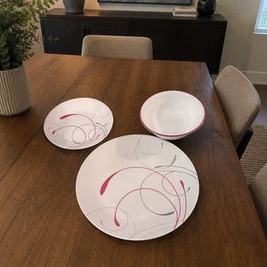 May include: A set of white dinnerware consisting of a large dinner plate, a smaller salad plate, and a deep bowl. Each piece features a decorative pattern of thin, flowing gray lines and vibrant pink teardrop-shaped accents across the surface.