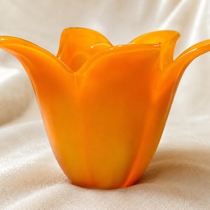 May include: A vibrant orange glass vase shaped like an open flower blossom. The vessel features smooth, curved edges that flare outward at the top, creating a wide, petal-like rim. The glass has a translucent quality with a glossy finish.