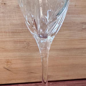 May include: A clear, stemmed wine glass with a decorative etched pattern on the bowl. The glass has a long, slender stem and a circular base. The background is a wooden surface.