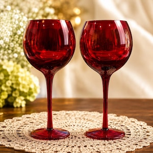 May include: Two matching wine glasses crafted from deep red translucent glass. Each glass features a wide, rounded bowl, a slender stem, and a circular base. The glass material is smooth and reflective, showing light highlights on the surface.