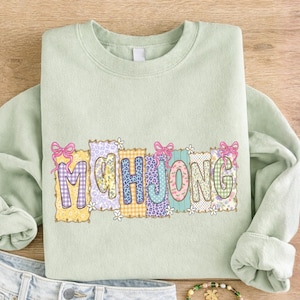 Könnte beinhalten: Ein hellgrünes Sweatshirt mit dem Wort "MAHJONG" in bunten, gemusterten Buchstaben. Jeder Buchstabe hat ein einzigartiges Design, darunter Vichy-, Blumen- und Leopardenmuster, mit rosa Schleifen und kleinen weißen Blumen. Das Sweatshirt liegt auf einer Holzoberfläche.