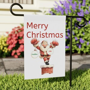 May include: A white garden flag with the words "Merry Christmas" in red. The flag features a cartoon Santa Claus holding ornaments. The ornaments have the names "Jonathan" and "Julie" on them. The flag is displayed on a black metal stand.