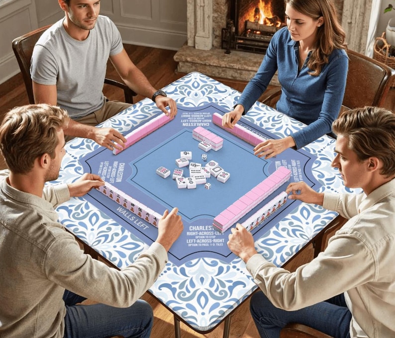 May include: Four people playing Mahjong around a square table. The table has a blue and white patterned surface with a central blue area. Pink tile racks and white tiles are visible. Text on the table reads "CHARLESTON" and "WALLS LEFT".