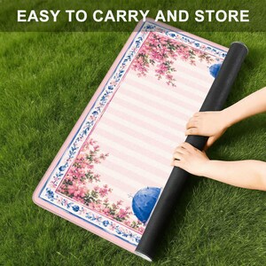 Pode incluir: Um tapete retangular com um centro de riscas rosa claro e brancas, bordado por um padr&atilde;o floral de flores cor-de-rosa e detalhes azuis. O texto EASY TO CARRY AND STORE est&aacute; impresso no topo. O tapete tem uma base preta e cantos arredondados.