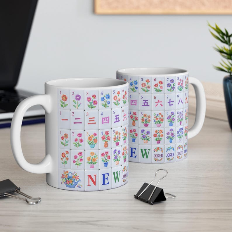 May include: Two white ceramic mugs with a floral tile design. The tiles feature colorful flowers and numbers, with the word "NEW" on one mug. The design is reminiscent of a game, with the word "JOKER" also visible.