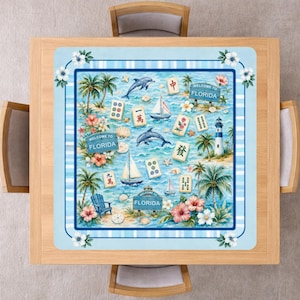 Puede incluir: Un mantel cuadrado con un tema de Florida. El dise&ntilde;o presenta palmeras, delfines, veleros y fichas de Mahjong. Las palabras "WELCOME TO FLORIDA" se muestran en letreros. El borde es azul claro con flores blancas.
