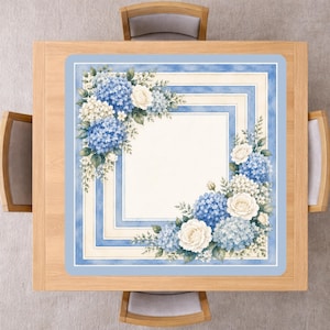Op de afbeelding: Een vierkante tafelloper met een bloemenontwerp. De loper heeft een lichtblauwe rand met een vierkant kaderontwerp. De hoeken zijn versierd met blauwe hortensia's, witte rozen en groene bladeren. De loper ligt op een houten tafel.