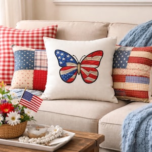 May include: A collection of decorative throw pillows on a beige sofa. One pillow features a butterfly design with the American flag colors. Other pillows have patchwork patterns and a flag design. A small American flag is in a basket of flowers.