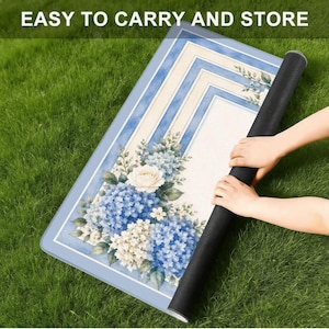 Op de afbeelding: Een opgerolde rechthoekige mat met een bloemenontwerp met blauwe hortensia's en witte rozen. De mat heeft een lichtblauwe rand en een cr&egrave;mekleurig midden met blauwe strepen. De tekst "EASY TO CARRY AND STORE" staat bovenaan.