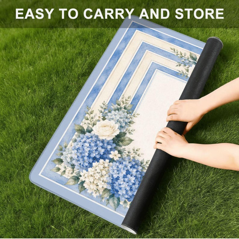 Op de afbeelding: Een opgerolde rechthoekige mat met een bloemenontwerp met blauwe hortensia's en witte rozen. De mat heeft een lichtblauwe rand en een cr&egrave;mekleurig midden met blauwe strepen. De tekst "EASY TO CARRY AND STORE" staat bovenaan.
