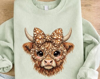 May include: A light green sweatshirt featuring a whimsical illustration of a Highland cow. The cow has brown fur, a polka-dot bow, heart-shaped glasses, and horns. The sweatshirt is made of a soft material.