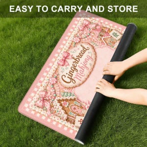 May include: A rectangular mat featuring a pink gingerbread house theme with the text "Gingerbread Mahjong" in a decorative oval frame, accented by pink bows, candy details, and a checkered border, with a black underside and rolled edge.