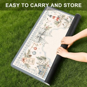 Op de afbeelding: Een opgerolde decoratieve mat met een winters tafereel met een vuurtoren, huizen en een grote strik. De mat heeft een blauw-witte geruite rand en de tekst "EASY TO CARRY AND STORE". De mat ligt op groen gras.