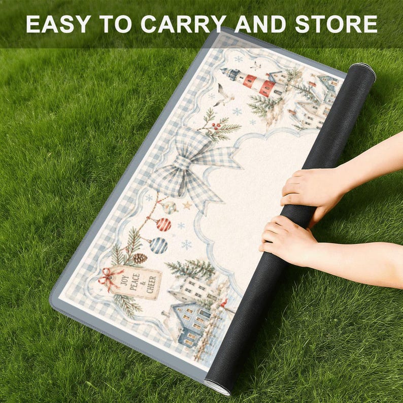 Op de afbeelding: Een opgerolde decoratieve mat met een winters tafereel met een vuurtoren, huizen en een grote strik. De mat heeft een blauw-witte geruite rand en de tekst "EASY TO CARRY AND STORE". De mat ligt op groen gras.