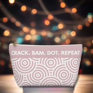 May include: A pink cosmetic bag with a gold zipper and the text "CRACK. BAM. DOT. REPEAT" in white. The bottom half of the bag features a white and pink concentric circle pattern. The background is blurred with bokeh lights.