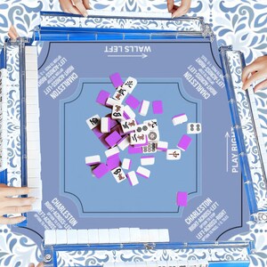 May include: A light blue Mahjong game set with white and purple tiles. The game board has text that reads "Walls Left" and "Play Right". The game is set on a patterned surface, with hands visible around the board.