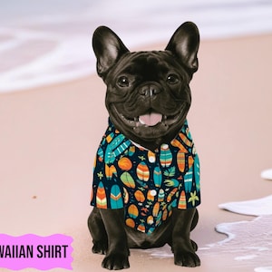 May include: A black French Bulldog wearing a Hawaiian shirt. The shirt is navy blue with a pattern of surfboards, flowers, and other tropical elements in shades of orange, teal, and yellow. The dog is sitting on a sandy beach.