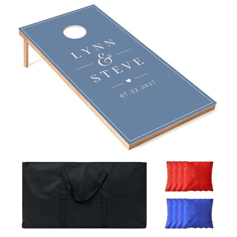 May include: A blue cornhole board with a white border and a circular hole, featuring the white text LYNN & STEVE, a small heart, and the date 07.22.2027. Includes a black carrying bag, four red bean bags, and four blue bean bags.