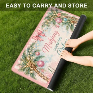 May include: A pink and white Mahjong mat with a palm tree design, Christmas lights, and the text "Mahjong Under the Palms." The mat is rolled up, showing a black underside. The text "EASY TO CARRY AND STORE" is at the top.
