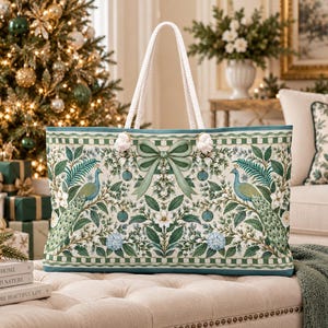 May include: A rectangular tote bag featuring a cream-colored background with a repeating pattern of green peacocks, white flowers, blue hydrangeas, and a central sage green bow. The bag has two thick white rope handles attached with metal grommets.