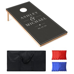 May include: A black cornhole board with a white border, featuring the printed text ASHLEY & MICHAEL, a small heart icon, and the date 06.25.26. The set includes a black carrying bag, four red bean bags, and four blue bean bags for the game.
