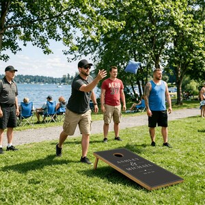 May include: A black cornhole board with a wooden frame featuring the white printed text Ashley & Michael and a date. The board has a circular hole at the top center and is positioned on a grassy lawn during an outdoor game session.