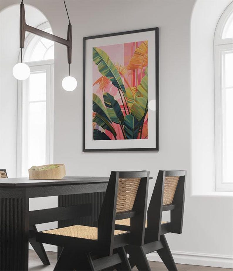 May include: A framed botanical print with a vibrant tropical plant design in shades of green, yellow, and pink. The artwork hangs above a black dining table and chairs with woven backs. The room is illuminated by hanging globe lights.