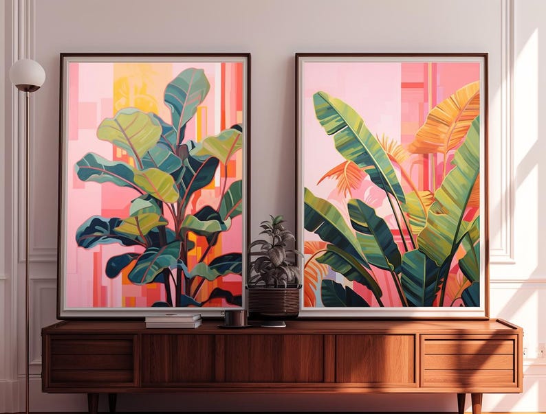 May include: Two framed botanical art prints. One features a large plant with green and blue leaves against a pink and orange background. The other print shows large green and yellow leaves with a pink and orange background. The prints are displayed on a wooden cabinet.
