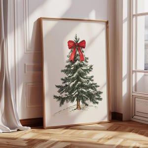 May include: A framed print featuring a detailed illustration of a Christmas tree adorned with a large red bow. The tree is green with a brown trunk, set against a neutral background. The artwork is in a gold frame, leaning against a wall.