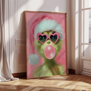 Puede incluir: Obra de arte enmarcada que presenta una criatura verde con gafas de sol rosas en forma de corazón y un gorro de Papá Noel, soplando una burbuja rosa. El fondo es de color rosa y el marco es dorado.