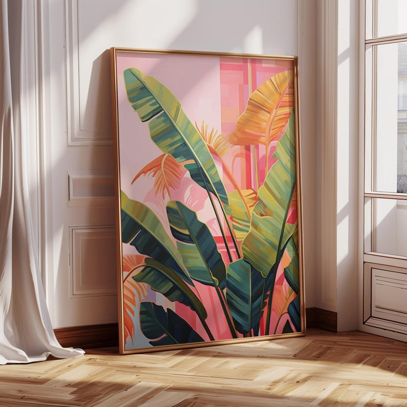 May include: A framed botanical art print featuring large, stylized banana leaves in shades of green, yellow, and orange against a pink and peach background. The artwork has a gold frame and leans against a white wall, creating a tropical theme.