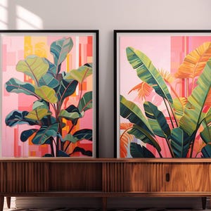 May include: Two framed botanical art prints. One features a large plant with green and yellow leaves against a pink and orange background. The other print shows large green and orange leaves against a pink background. The frames are black.