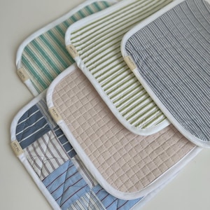 May include: Set of four quilted hot pads with white piping. The hot pads are made of different patterned fabrics, including blue and white stripes, green and white stripes, and a patchwork design. Each hot pad has a white label that says "HOT PAD".