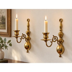May include: A pair of polished brass wall sconces, each featuring a vertical mounting plate with a decorative finial, a curved arm, and a circular candle cup. Each sconce holds a single off-white taper candle with a lit flame, creating a warm glow.