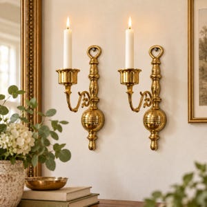 May include: A pair of polished brass wall sconces, each featuring a vertical mounting plate with a decorative finial, a curved arm, and a circular candle cup. Each sconce holds a single white taper candle, creating a symmetrical display.