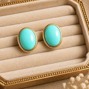 May include: Two turquoise oval stud earrings with a gold-coloured border. The earrings are presented in a gold-trimmed jewellery box with a beige interior. The earrings have a smooth, polished finish.