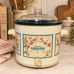 May include: A cream-colored Rival Crock-Pot slow cooker with a clear glass lid and two side handles. The exterior features a floral pattern in teal frames with the text Rival Crock-Pot Slow Cooker/Server and settings for Off, Low, and High.
