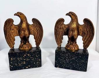 May include: Two bronze-colored eagle sculptures with outstretched wings. Each eagle sits atop a black rectangular base with gold speckles. The sculptures are detailed and decorative, suitable for home decor.