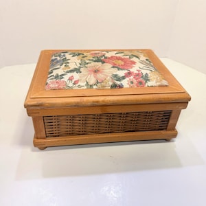 May include: A rectangular wooden sewing box featuring a light brown finish and a woven wicker side panel. The lid is padded and upholstered with a cream-colored fabric displaying a vibrant floral pattern in shades of pink, red, and blue.