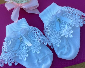 May include: A pair of white baby socks with delicate white lace trim. Each sock features a satin ribbon and a small silver cross charm. The socks are displayed on a pink surface, with a wicker basket and a pink bow in the background.