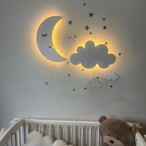 May include: A white, illuminated moon and cloud wall decor with a warm yellow glow. The moon has a smiling face and closed eyes. Additional gray star and cloud decals are on the wall. A white crib with cloud-shaped pillows and a teddy bear are in the foreground.