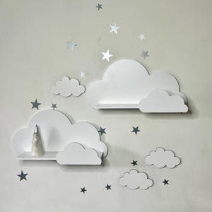 Peut inclure: Étagères blanches en forme de nuage avec une petite figurine sur l'une d'elles et des décorations en forme d'étoiles argentées. Les étagères sont fixées sur un mur de couleur claire, créant un affichage décoratif et fantaisiste.