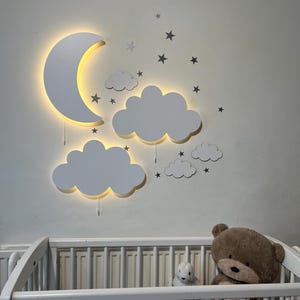 May include: White wooden moon and cloud night light with a warm glow. The light is mounted on a white wall above a white cot with a teddy bear and a stuffed animal inside.