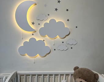 May include: White wooden moon and cloud night light with a warm glow. The light is mounted on a white wall above a white crib with a teddy bear and a stuffed animal inside.