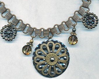 May include: A silver and gold necklace with a large, ornate pendant featuring a flower design. The necklace also has several smaller, dangling charms, including a round, iridescent bead and a cluster of black beads.