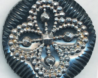 May include: A black button with a silver design of a four-leaf clover. The button has a textured surface.